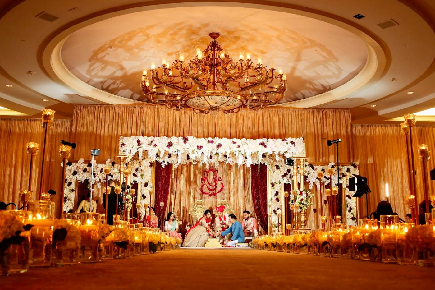 A wedding ceremony under a floral mandap, with guests seated along a candlelit aisle.