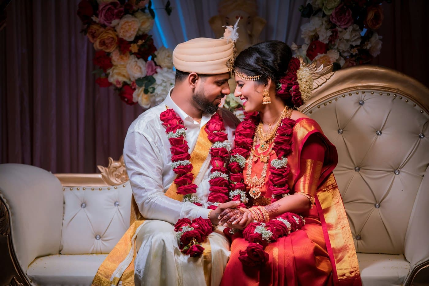 A bride and groom in red and white sit together on a sofa, holding hands and smiling.