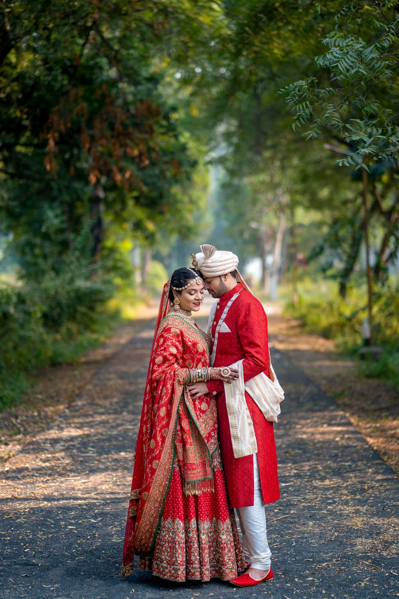 A bride and groom in red wedding clothes stand close together on a tree-lined path.